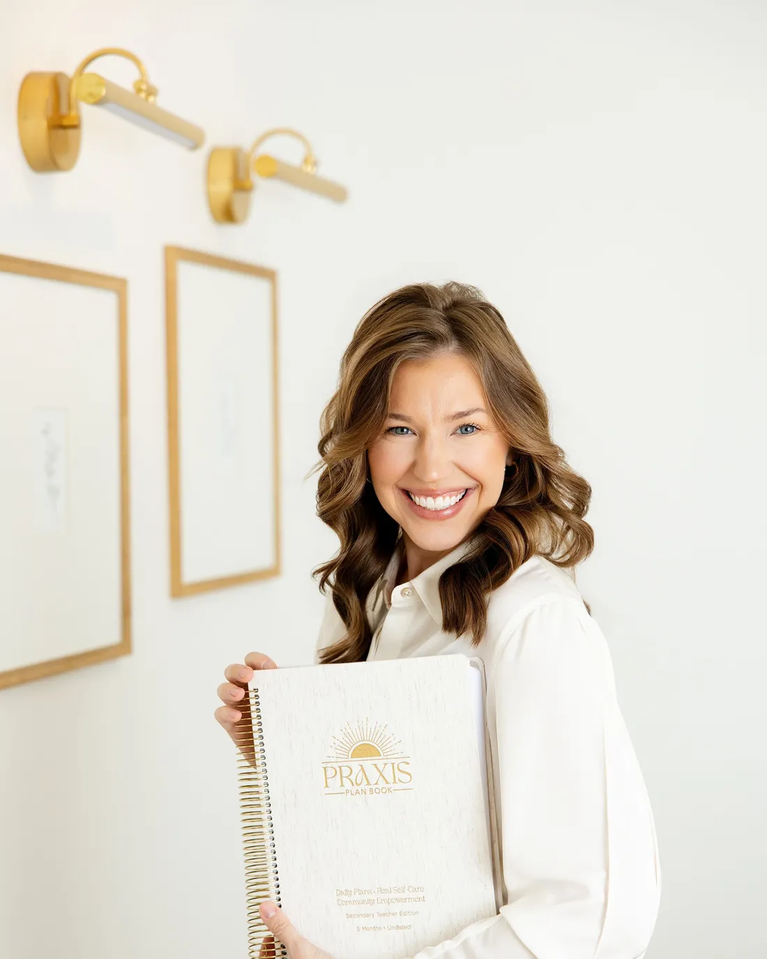 Sarah Bland standing with the Praxis Plan Book in her arms, gold picture lights on the wall behind her