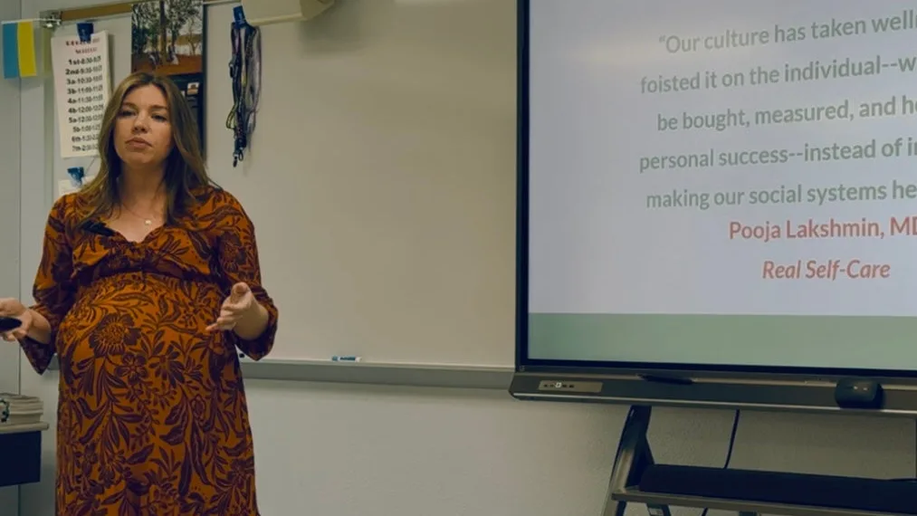 Sarah Bland leading a staff wellbeing session in a classroom, mid-sentence beside a screen showing a Pooja Lakshmin quote from Real Self-Care